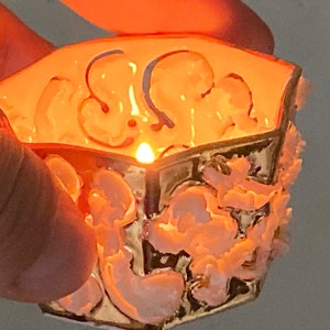 May include: A small, octagonal, ceramic candle holder with a white and gold design. The holder has a raised, floral pattern and is lit by a candle inside.