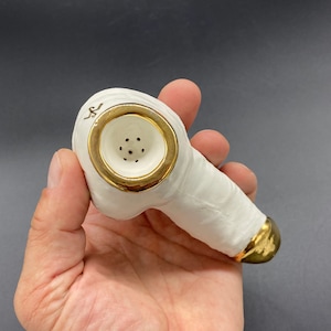 May include: White ceramic pipe with gold accents. The pipe has a round bowl with a gold rim and a small hole in the center. The pipe is held in a hand.