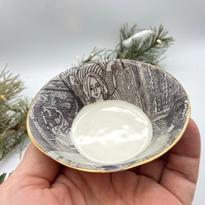 Pode incluir: Uma pequena tigela de cerâmica branca com uma borda dourada. A tigela é decorada com uma ilustração em preto e branco de uma mulher com pássaros e outros desenhos intrincados.