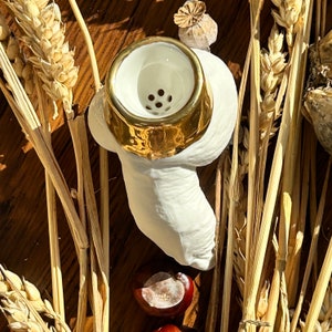 May include: White ceramic pipe with a gold rim and a small hole in the center. The pipe is shaped like a curved tube and is sitting on a bed of dried wheat stalks.