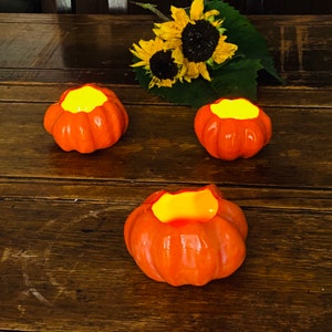 Puede incluir: Tres portavelas de cerámica naranja en forma de calabaza con un cálido resplandor de las velas en su interior. Los portavelas están sobre una superficie de madera.