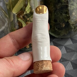 May include: A white ceramic finger with a gold top and a cork base. The finger is a decorative object.