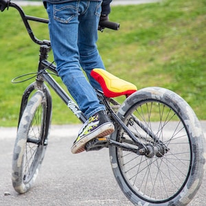 May include: A person riding a black BMX bike with a yellow and red seat. The bike has a black frame and black tires with a camouflage pattern.