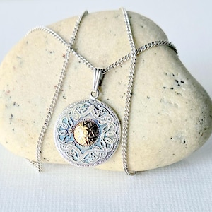May include: A silver-toned pendant necklace with a circular design. The pendant features an ornate pattern with a gold-colored center. The necklace is displayed on a light-colored stone against a white background. The chain is delicate and silver.