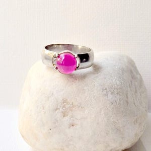 May include: A silver ring with a large, round, bright pink gemstone. The ring is set on a white stone against a white background. The band of the ring is thick and smooth.