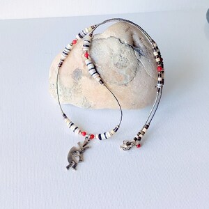 May include: A handcrafted necklace featuring a silver pendant of a Kokopelli figure. The necklace has a delicate wire design with alternating white, black, and brown beads, accented by small red beads. The necklace is displayed on a light-colored stone.