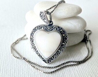 May include: A heart-shaped locket pendant featuring a central white mother-of-pearl inlay surrounded by a dark silver frame with intricate marcasite stone detailing. The pendant hangs from a thin, matching silver box chain necklace.