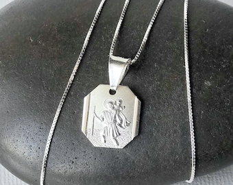 May include: A silver-toned pendant necklace featuring an octagonal charm with an engraved depiction of a figure carrying a child on their shoulder. The pendant hangs from a delicate silver-toned box chain, showcasing a polished metallic finish.