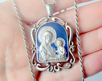May include: A silver-toned pendant necklace featuring a rectangular icon with a deep blue enamel background. The pendant depicts a raised silver relief of a figure holding a child, surrounded by an ornate, scrolling silver frame and a rope chain.