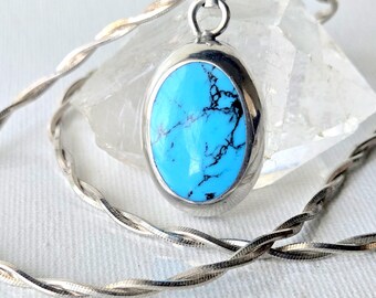 May include: An oval turquoise pendant with black matrix veining set in a polished silver bezel. The pendant hangs from a silver twisted rope chain, featuring a smooth, rounded metal frame that secures the vibrant blue stone in place.