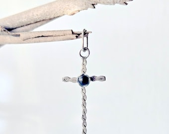 May include: A silver-toned metal cross pendant featuring a twisted rope-like texture on the vertical and horizontal bars. A small, round, dark blue stone is set at the center where the bars intersect, topped with a small circular jump ring.