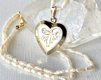 May include: A gold heart-shaped locket pendant featuring an engraved floral pattern on the front, suspended from a necklace composed of small, irregular white freshwater pearls with a gold-toned metal clasp and connector hardware.