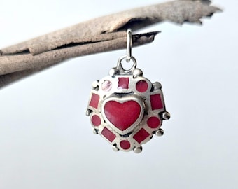 May include: A silver pendant featuring a central red heart-shaped inlay surrounded by smaller red geometric shapes, including squares and circles, set within a silver frame with a small jump ring at the top for attachment to a chain or cord.