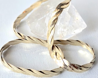 May include: A gold-toned herringbone chain bracelet featuring a twisted, braided design. The metal surface has a reflective, polished finish with fine, diagonal etched detailing throughout the links, creating a textured appearance.