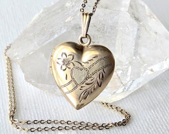 May include: A gold-toned heart-shaped locket pendant suspended from a delicate matching chain. The locket features an engraved floral motif and a smaller heart outline on its front surface, showcasing a polished metallic finish throughout.