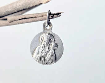 May include: A circular silver-toned metal pendant featuring a raised relief design of a figure with a halo holding a smaller figure. The pendant has a small loop at the top for attachment and displays a matte, textured finish on its surface.