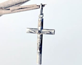 May include: A silver-toned metal crucifix pendant featuring a central figure of Jesus on the cross. The cross has a smooth, cylindrical bar design with a small rectangular plaque at the top. A metal jump ring is attached to the top for suspension.