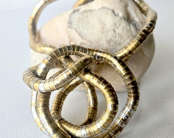 May include: A flexible, segmented metal chain necklace featuring alternating gold and silver-toned cylindrical links. The chain is coiled into a series of loops, creating a continuous, fluid, and interconnected circular pattern with a metallic finish.