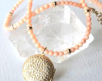 May include: A round gold-tone metal locket featuring an embossed floral pattern on the front, suspended from a beaded necklace. The necklace consists of small, round, light peach-colored beads interspersed with polished gold-tone metal spacer beads.