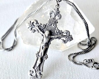 May include: A silver-toned metal crucifix pendant featuring an ornate, scrolled design and a central figure of Jesus. The cross includes the letters INRI at the top and hangs from a matching silver-toned box chain necklace with a circular clasp.
