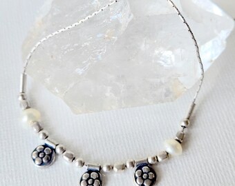 May include: A delicate silver chain necklace featuring three small circular charms with a raised flower petal design, interspersed with smooth round silver beads and two small white pearls, finished with a silver clasp and a small rectangular tag.