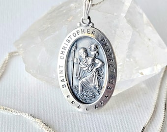 May include: A silver oval pendant necklace featuring a relief of Saint Christopher carrying a child. The border is inscribed with the text SAINT CHRISTOPHER PROTECT US and decorated with small stars. The pendant hangs on a thin silver box chain.