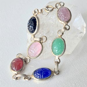 May include: A silver link bracelet featuring six oval-shaped, carved scarab gemstones in various colors including dark blue, light pink, deep red, bright blue, green, and pale purple, connected by small silver metal rings and a circular clasp.