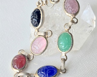 May include: A silver link bracelet featuring six oval-shaped, carved scarab gemstones in various colors including dark blue, light pink, deep red, bright blue, green, and pale purple, connected by small silver metal rings and a circular clasp.