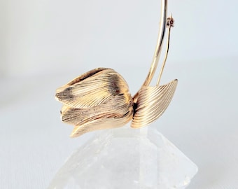 May include: A gold-toned metal brooch shaped like a tulip flower with a textured, ribbed surface on the petals and a single curved stem. The brooch features a pin mechanism on the back and is displayed resting on a clear, faceted crystal base.