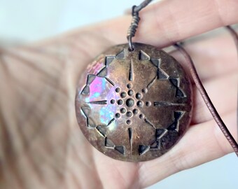 May include: A circular copper pendant featuring a geometric starburst pattern with etched lines and small circular indentations. The metal surface displays a weathered patina with iridescent purple and blue highlights, suspended from a thin brown cord.