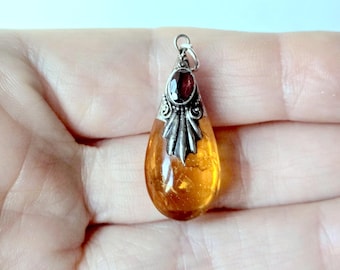 May include: A teardrop-shaped amber pendant featuring a polished golden-orange stone set in a silver metal cap. The cap is decorated with a small, oval-cut dark red garnet gemstone and includes a silver jump ring at the top for attachment.