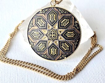 May include: A round pendant necklace featuring a black and gold geometric pattern with a central square motif surrounded by eight pointed shapes. The pendant is suspended from a gold-toned metal chain and has a thin gold-colored metal rim.