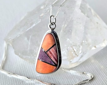 May include: A silver pendant necklace featuring a teardrop-shaped inlay of orange and purple stone segments separated by thin silver lines. The pendant hangs from a delicate silver box chain, showcasing a geometric mosaic design with smooth edges.