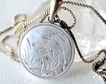 May include: A round silver locket pendant featuring an engraved heron standing among reeds and a palm tree. The pendant is attached to a silver box chain necklace with a polished bail, showcasing detailed metalwork and a metallic finish.