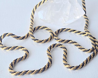 May include: A twisted rope chain necklace featuring alternating strands of metallic gold and dark charcoal black cord. The necklace includes a gold-toned metal clasp mechanism at the top, connecting the two ends of the twisted cord design.