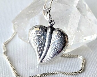 May include: A silver heart-shaped locket pendant featuring an engraved leaf-like pattern on both sides, suspended from a delicate silver box chain. The locket is positioned in front of a clear, multifaceted quartz crystal stone.