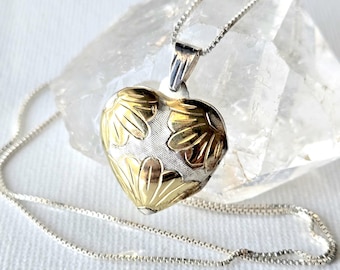 May include: A heart-shaped pendant necklace featuring a two-tone design with gold-colored floral accents on a textured silver-colored background. The pendant hangs from a delicate silver-colored box chain, resting against a clear crystal.