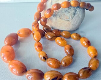 May include: A necklace composed of polished, oval-shaped beads in varying shades of translucent orange, honey, and deep amber. The beads feature natural swirling patterns and are strung together in a continuous loop, resting against a smooth, light-colored stone.