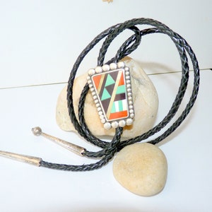 May include: A bolo tie with a geometric design. The rectangular pendant features a mosaic of colorful stones in shades of orange, green, black, and beige, set in a silver frame. The black braided leather cord has silver tips.