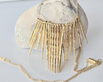 May include: A gold-toned necklace featuring a fringe-style pendant composed of multiple thin, hammered metal spikes of varying lengths suspended from a delicate chain. The metal surface has a textured, reflective finish throughout the design.