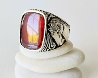 May include: A silver ring featuring a rectangular, smooth, deep red stone set in the center. The sides of the silver band are detailed with an embossed, sculpted horse head design, showing the profile of the animal with flowing mane textures.