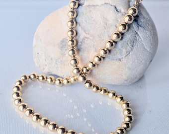 May include: A gold-toned beaded necklace featuring a continuous strand of polished, spherical beads. The necklace is draped over a smooth, light-colored, rounded stone, showcasing the reflective metallic surface and uniform size of each bead.