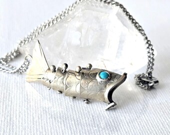 May include: A silver-toned articulated fish pendant necklace featuring a small, round, bright blue stone set as the eye. The fish body is composed of segmented metal plates with small spherical accents along the top and bottom edges.