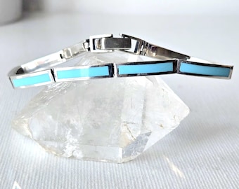 May include: A silver-toned metal bracelet featuring a series of rectangular links inlaid with smooth, bright turquoise-colored segments. The bracelet includes a secure metal clasp mechanism and is displayed resting on a clear, faceted crystal.