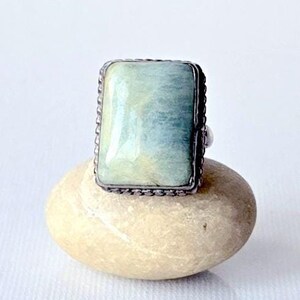 May include: A rectangular ring with a light blue and cream-colored stone. The stone is set in a silver-colored metal with a textured border. The ring is displayed on a small, light-colored stone against a white background.