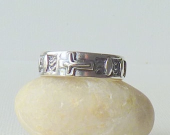 May include: A silver band ring featuring a repeating pattern of embossed crosses and oval shapes. The metal has a polished finish with darkened recessed areas that highlight the intricate details of the religious symbols and geometric motifs.