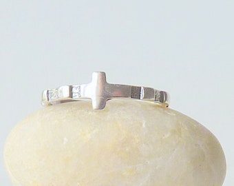 May include: A silver-toned ring featuring a central cross-shaped design element. The band is constructed with alternating smooth metallic segments and small, clear, square-cut stones embedded along the circumference of the ring.
