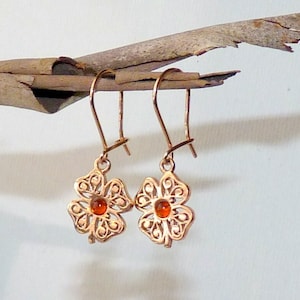 May include: A pair of copper-colored earrings. Each earring features a four-leaf clover design with an orange center stone. The earrings are suspended from a simple hook. The earrings are displayed on a wooden branch against a white background.