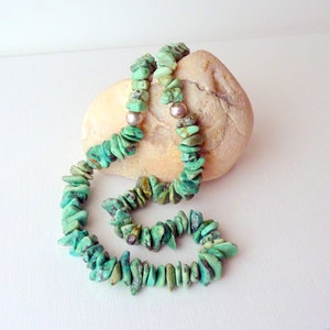 May include: A turquoise chip necklace with two silver beads. The necklace is draped over a light brown stone, set against a white background. The turquoise chips vary in size and shade, creating a natural, earthy aesthetic.
