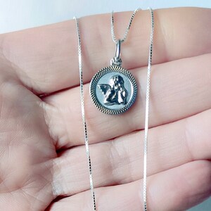 May include: A silver necklace featuring a round pendant with an angel design. The pendant has a detailed image of an angel within a textured border. The chain is a delicate, square-linked style. The necklace is displayed against a light background.